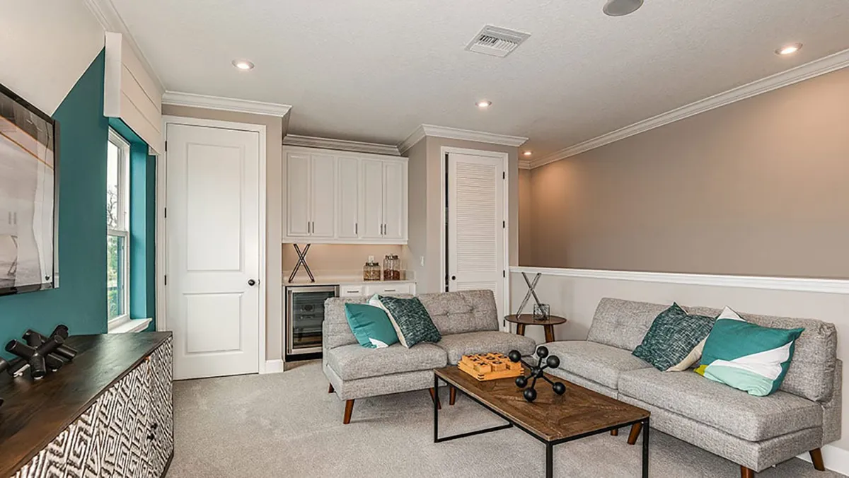 A modern living room with a sectional gray sofa, teal and white accent pillows, a wooden coffee table, a sideboard, wall art, built-in cabinets, a mini-fridge, and soft beige and teal wall colors.