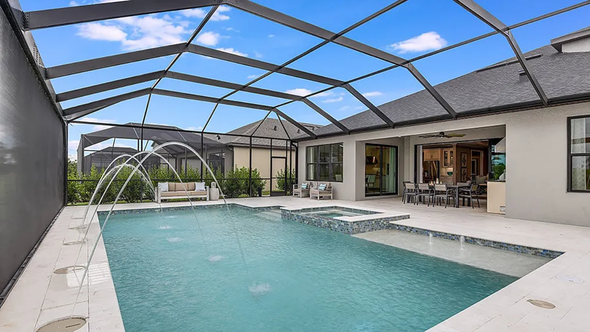 A modern screened-in backyard pool area with water jets, a hot tub, outdoor seating, and an open view into the house. The area features light stone flooring and a clear blue sky above.