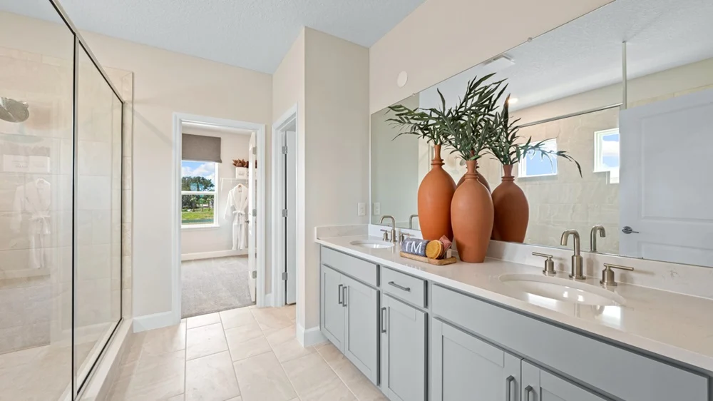 Modern bathroom with a double-sink vanity, large mirror, three orange vases with greenery, light gray cabinets, a glass shower enclosure, and a window letting in natural light.