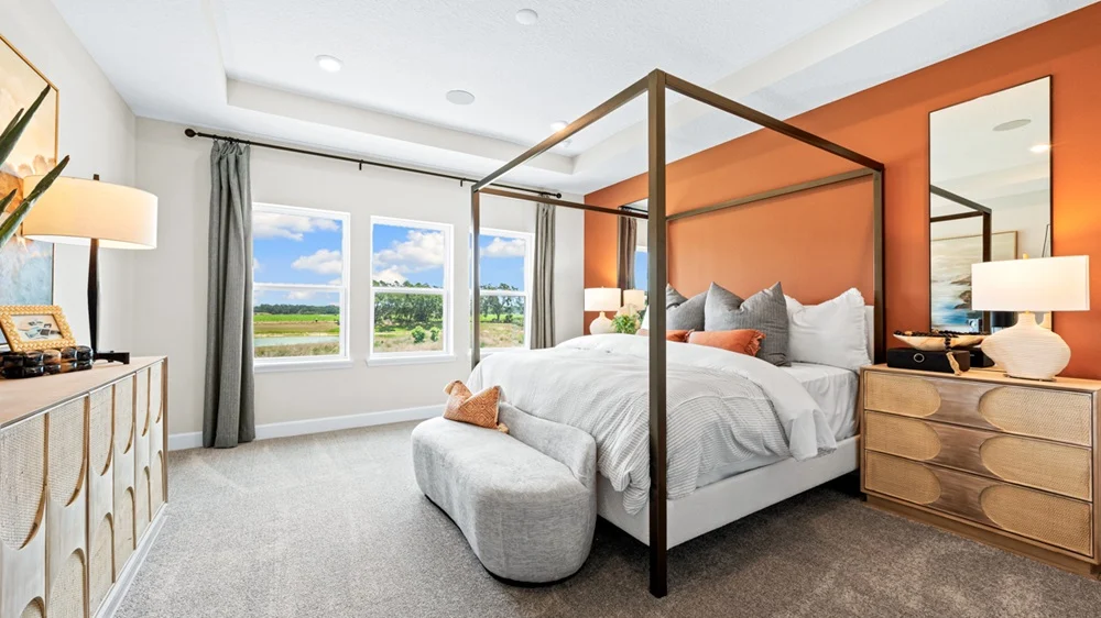 A modern bedroom with a four-poster bed, white bedding, orange accent wall, large windows with a scenic view, light wood furniture, a grey ottoman, and contemporary decor.