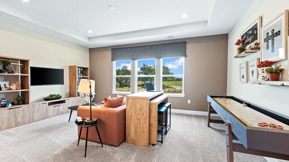 Bright, modern living room with a sofa, TV, shuffleboard table, bookshelves, wall art, and large windows overlooking greenery, decorated in neutral and wood tones.