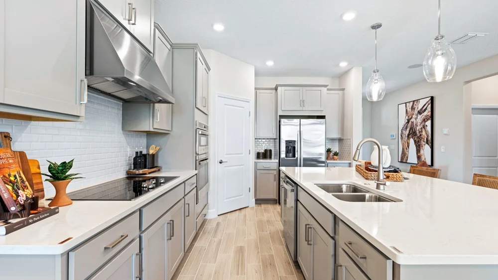 Modern kitchen with light gray cabinets, stainless steel appliances, double sink island, pendant lights, and wood-style flooring. A cookbook, utensils, and potted plant sit on the white countertops. Artwork hangs on the wall in the background.