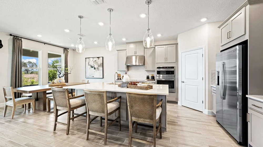 Modern kitchen with island seating, wicker chairs, stainless steel appliances, pendant lights, light cabinets, wood-look tile floors, and large windows letting in natural light. Dining table with chairs is visible on the left.