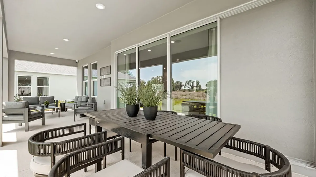 Modern covered patio with a sleek black dining table, six woven chairs, two potted plants on the table, and a seating area with armchairs in the background, all against large sliding glass doors.