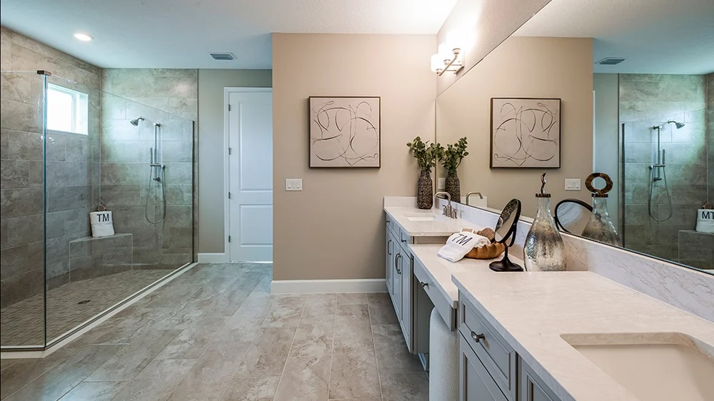 Modern bathroom with a large glass-enclosed shower, double vanity with white countertops, decorative vases, neutral tile floors, beige walls, and abstract art on the wall. A window provides natural light.