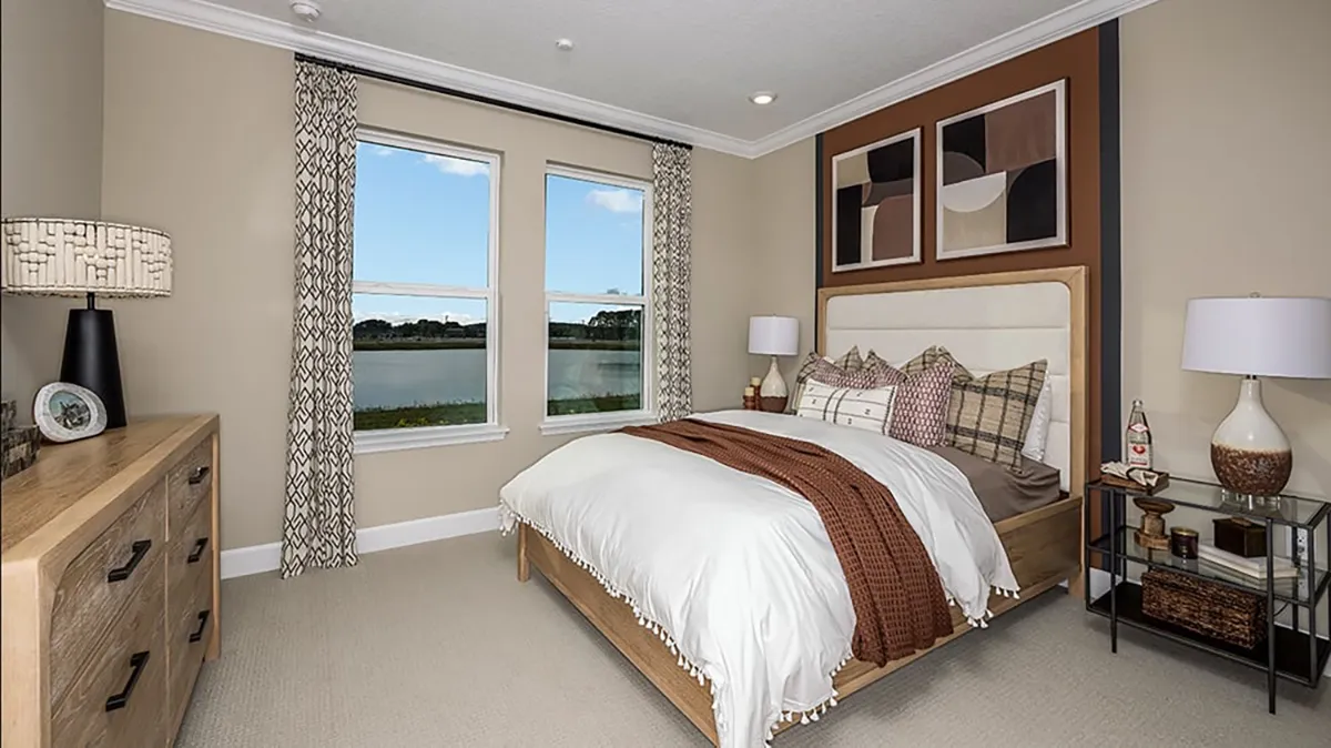 A modern bedroom with a wooden bed, white and plaid bedding, brown throw blanket, two windows with patterned curtains, abstract wall art, two bedside tables with lamps, and a wooden dresser.