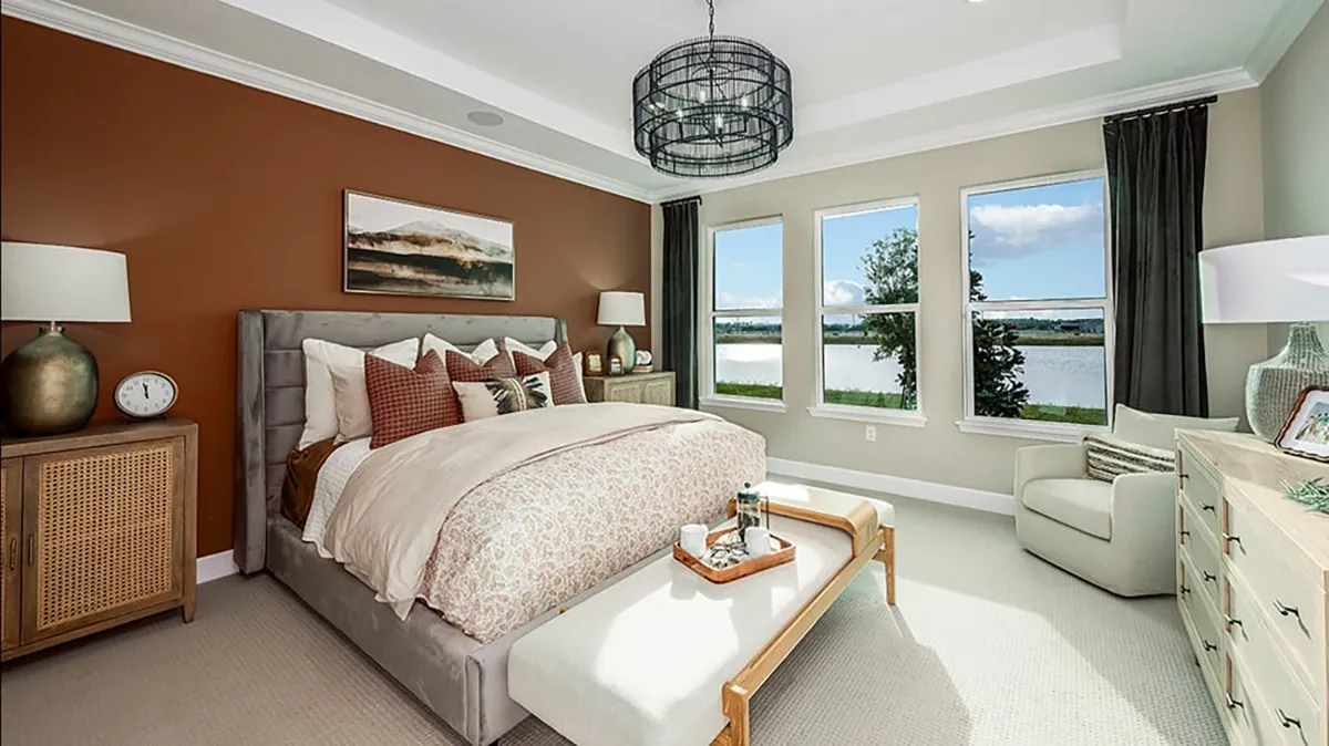 Modern bedroom with a large bed, brown accent wall, artwork above the headboard, two nightstands with lamps, bench at the foot of the bed, dresser, armchair, and three windows overlooking a lake and trees.