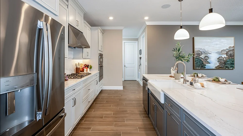 Modern kitchen with stainless steel appliances, white and gray cabinetry, marble countertops, pendant lighting, a kitchen island with a sink, wood-look tile flooring, and a landscape painting on the wall.