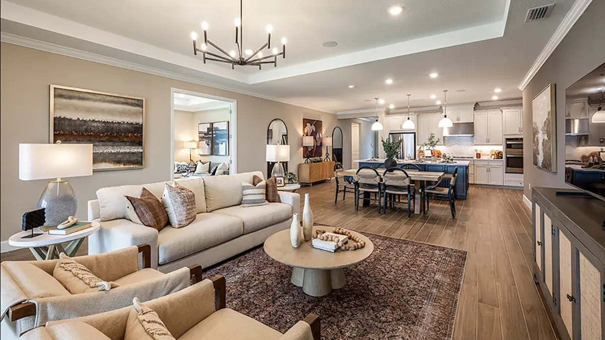 Open-concept living room and kitchen with beige sofas, armchairs, a round coffee table, dining table, and modern lighting. The kitchen features white cabinets, a large island, and pendant lights.