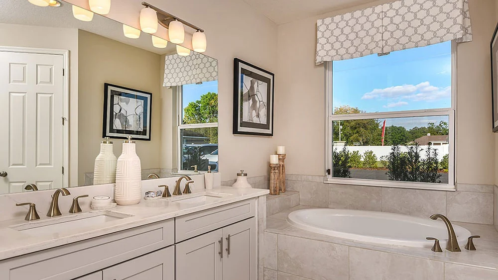 Modern bathroom with a double-sink vanity, large mirror, two white decorative bottles, a soaking tub, a window with a geometric valance, and framed wall art above beige tiled walls.