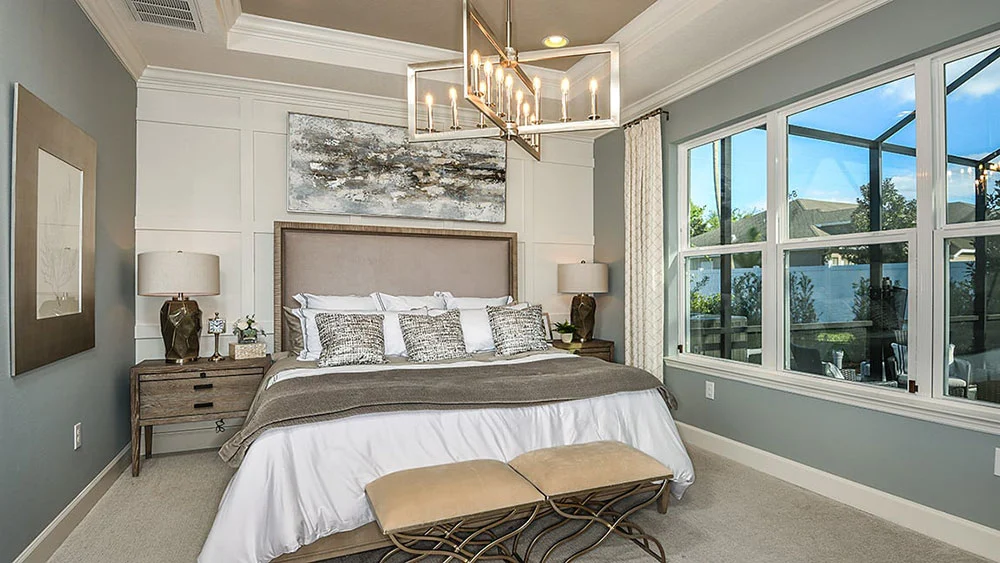 Modern bedroom with a large bed, gray and white bedding, two nightstands with lamps, abstract wall art, a chandelier, large windows with outside view, and a bench at the foot of the bed.