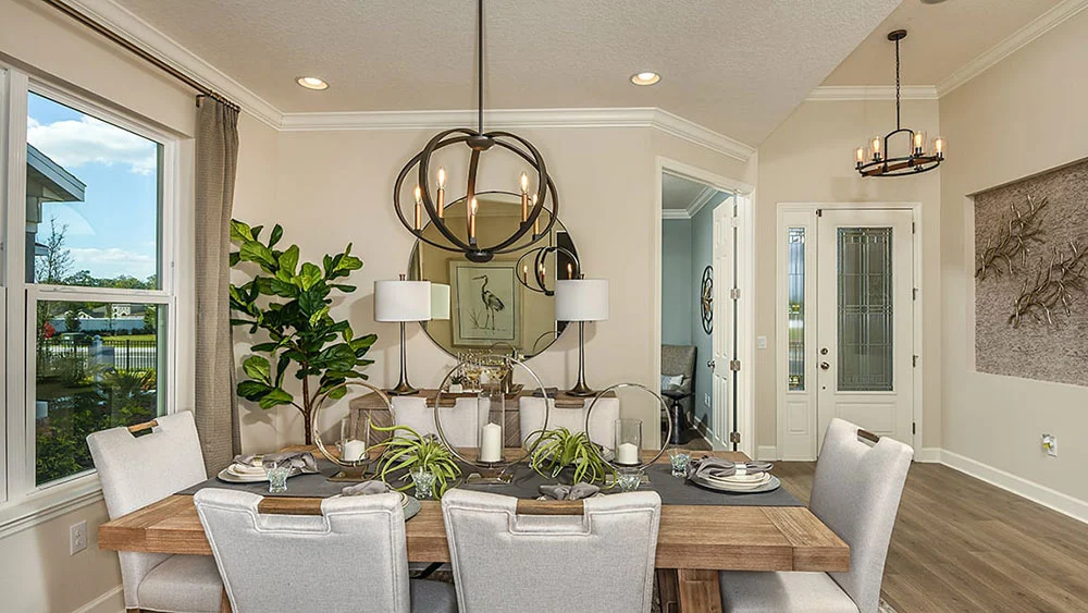 A modern dining room with a wooden table set for six, beige upholstered chairs, a circular chandelier, large window, potted plant, wall art, and decorative accents in a bright, neutral space.