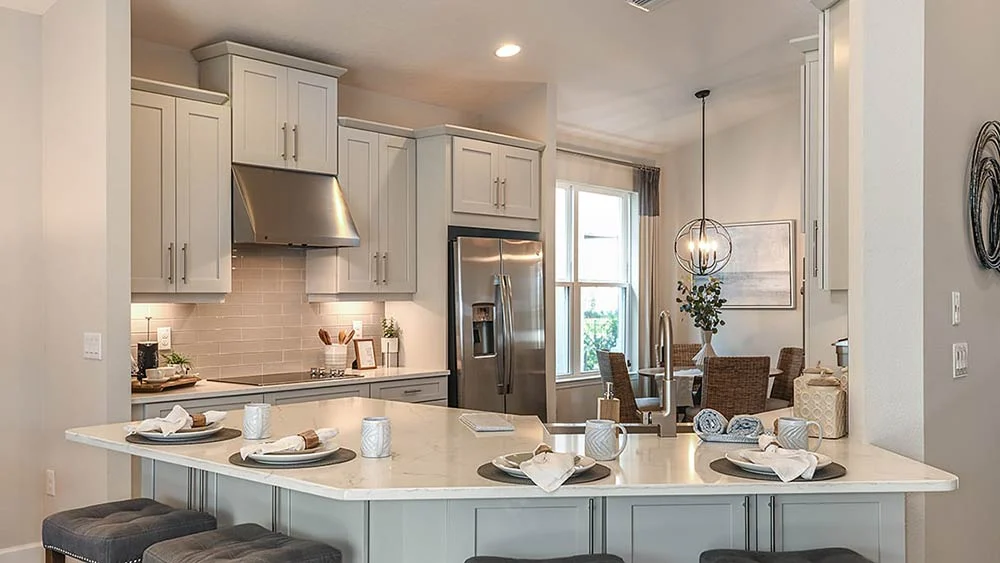 Modern kitchen with white cabinets, stainless steel appliances, and a large island set with four place settings and stools. In the background, a dining area with a round table and a hanging light fixture is visible.