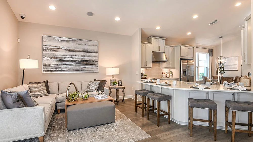 Modern open-concept living room and kitchen with neutral tones, a sectional sofa, coffee table, barstools at a white island, stainless steel appliances, and contemporary decor. Warm lighting creates a cozy atmosphere.