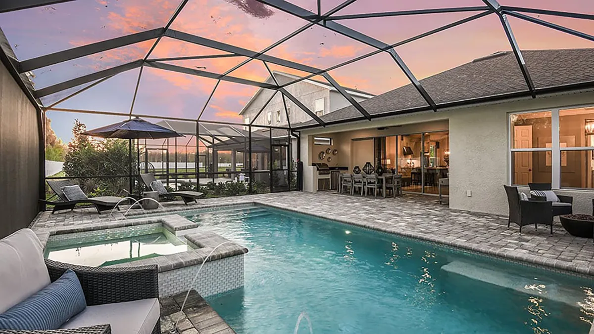 A screened-in backyard patio features a swimming pool with a spa, outdoor seating, and dining area. The sky is colorful at sunset, and the house’s windows reveal a warmly lit interior.