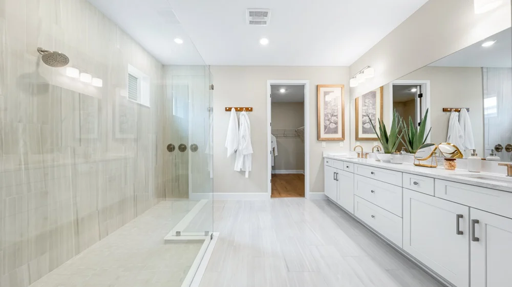 Modern bathroom with a large glass-enclosed shower, double sinks, white cabinets, two robes on wall hooks, decorative plants, and a walk-in closet in the background. The space is bright and neatly organized.