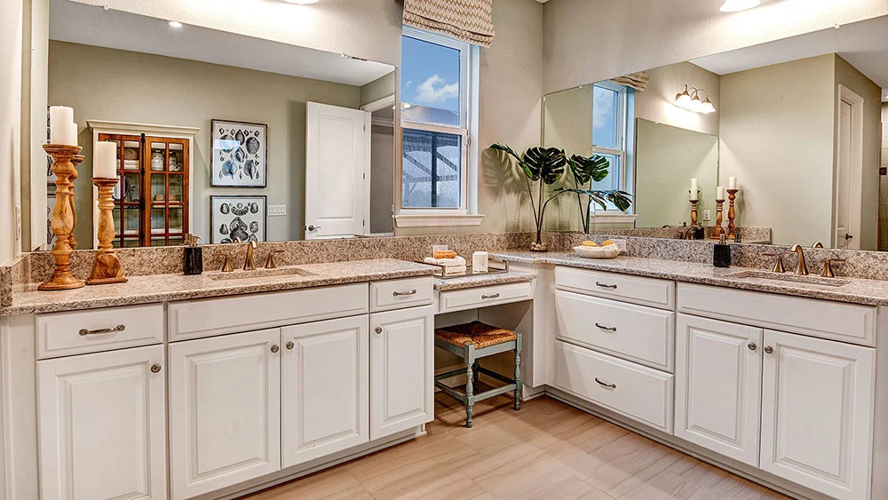 Modern bathroom with dual sinks, white cabinets, marble countertops, large mirrors, decorative candles, potted plant, and a small stool under the counter. A window lets in natural light above the sinks.