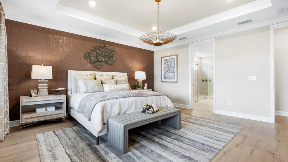 A modern bedroom with a large bed, decorative pillows, a textured brown accent wall, two nightstands with lamps, a gray bench at the foot of the bed, a striped rug, and a stylish light fixture.