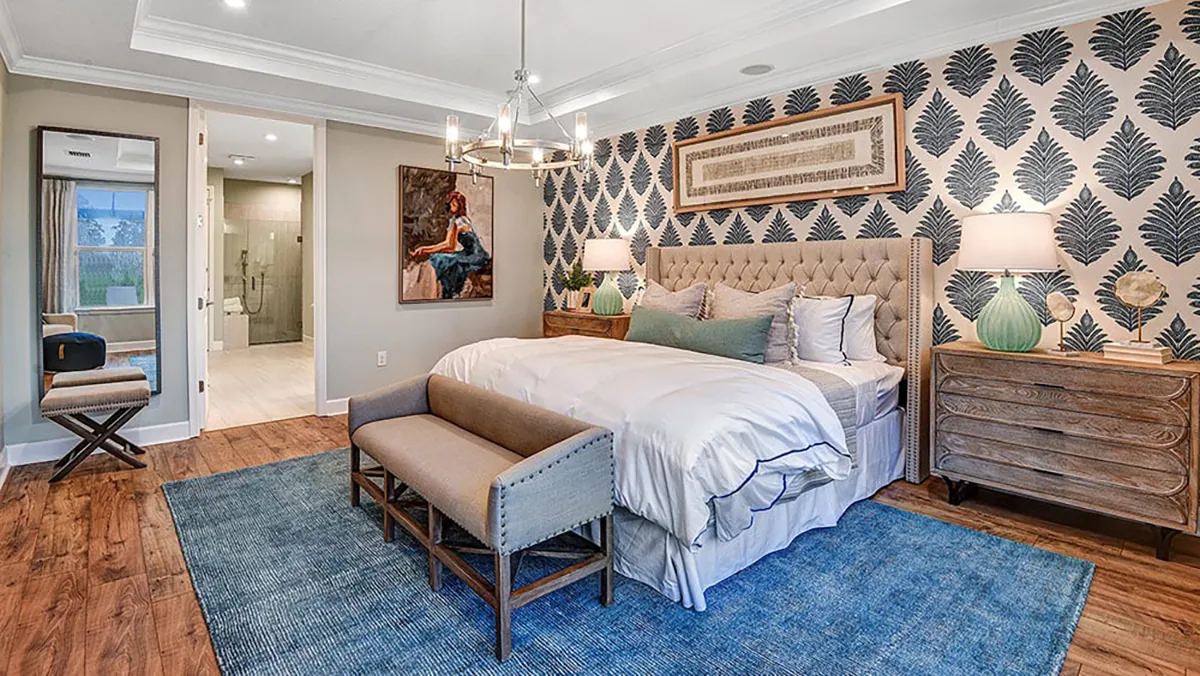 A stylish bedroom with a large bed, blue rug, bench, wooden dressers, two lamps, patterned wallpaper, modern chandelier, and artwork. An open door reveals a bathroom with a glass shower.