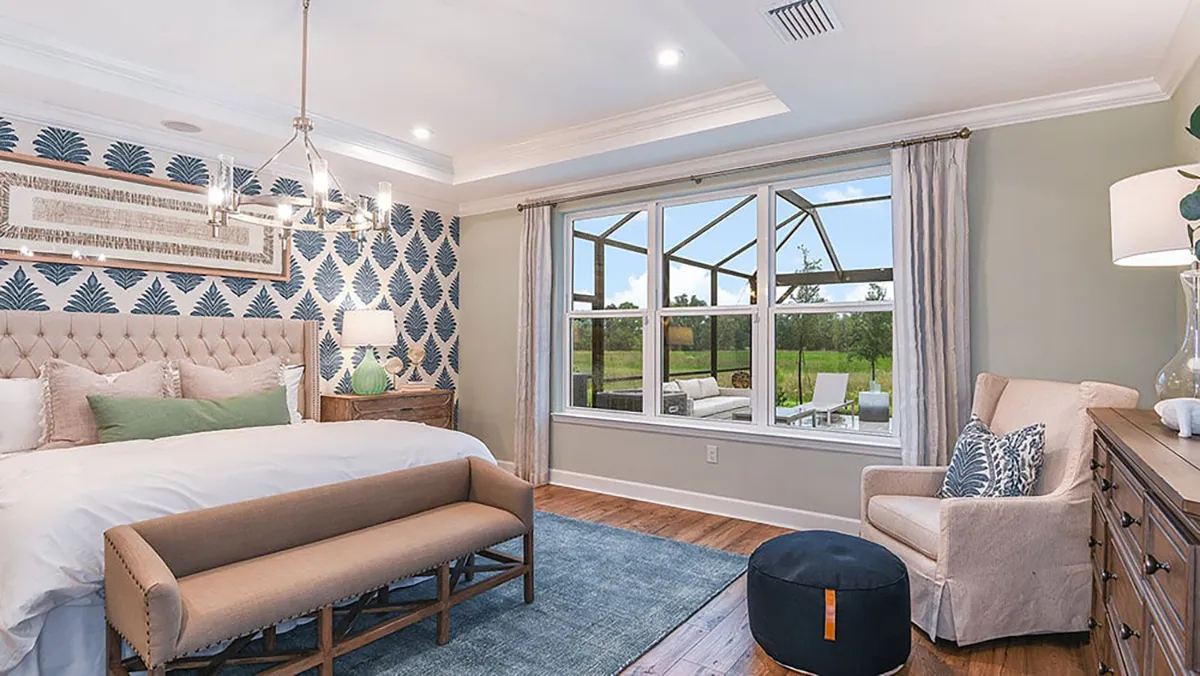 A bright, modern bedroom with a patterned accent wall, upholstered bed, bench, blue rug, armchair, large window with outdoor view, wood flooring, and neutral decor.