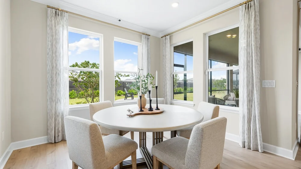 Bright dining area with a round white table, four upholstered chairs, a decorative centerpiece, and large windows allowing natural light and views of greenery outside. Light-colored curtains frame the windows.