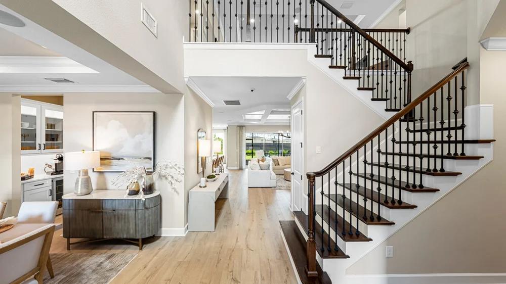 Modern home interior with wooden staircase, light wood floors, and open living space. White walls, a console table with decor, and artwork are visible. Natural light streams in from large windows in the background.