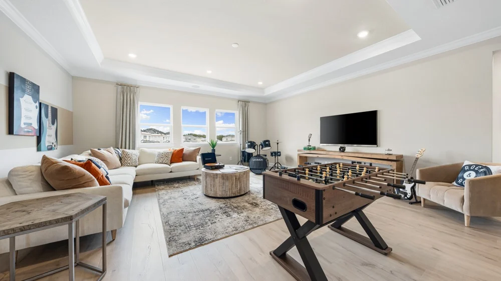 A spacious, modern living room features a beige sectional sofa with cushions, a round coffee table, a foosball table, wall art, a large TV, musical instruments, and three windows letting in natural light.