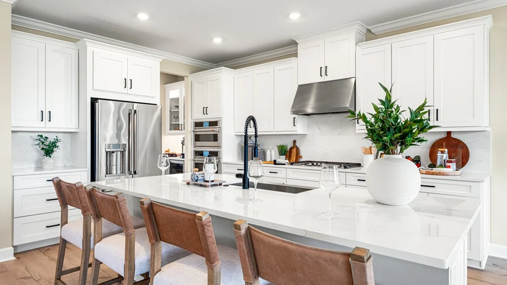Modern kitchen with white cabinets, stainless steel appliances, a large marble island with a black faucet, four brown leather barstools, and a white vase with green leaves on the countertop.