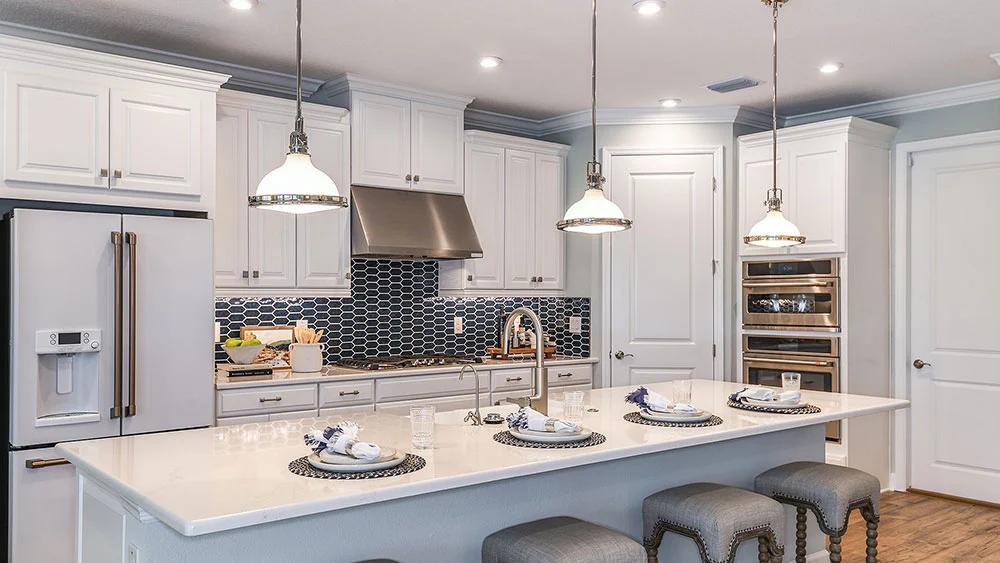 A modern kitchen with white cabinets, stainless steel appliances, a white island with sink, three gray stools, pendant lights, and a navy blue hexagon tile backsplash. Four place settings are arranged on the island.