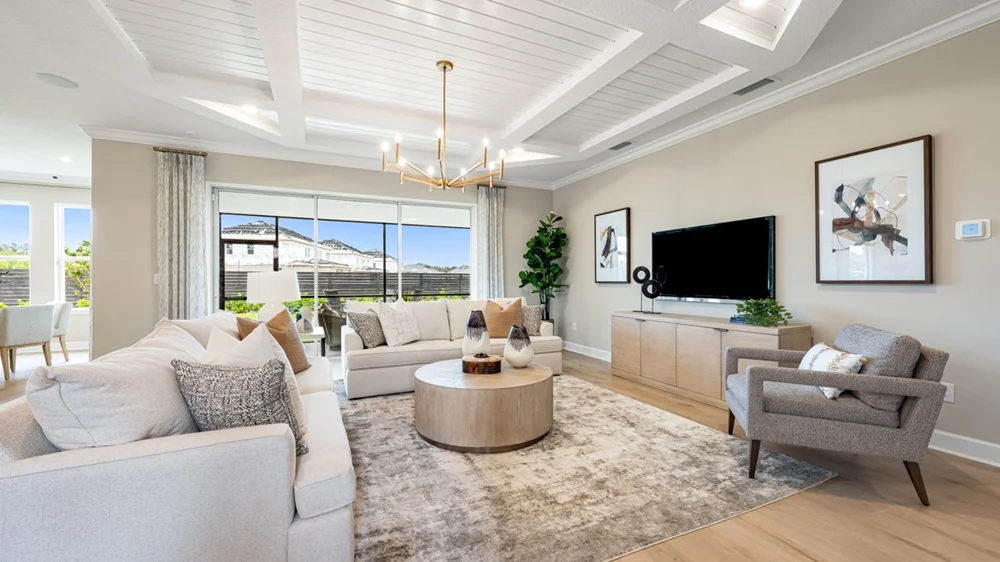 Modern living room with neutral tones features two sofas, a gray armchair, round coffee table, large rug, wall-mounted TV, framed art, indoor plant, chandelier, and large windows letting in natural light.