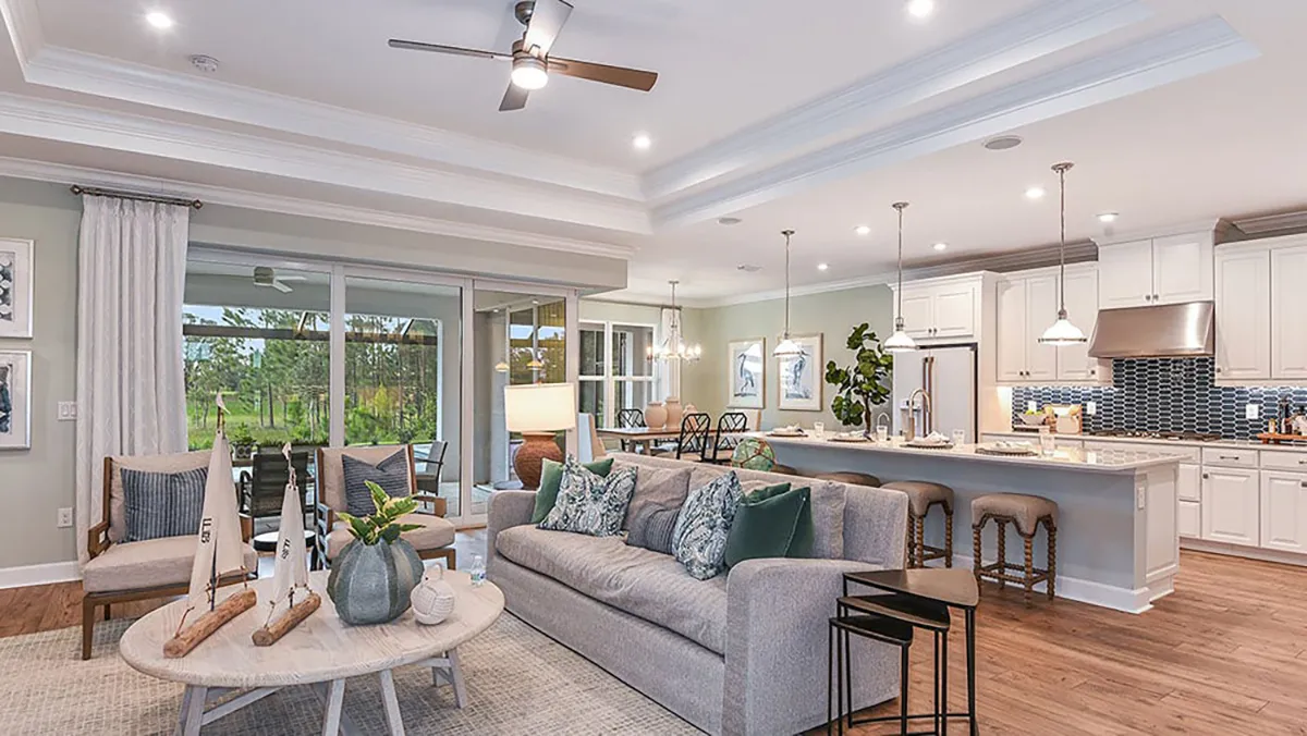 A modern open-concept living room and kitchen with a gray sofa, decorative pillows, round coffee table, bar stools at a kitchen island, and large sliding glass doors leading to a green outdoor view.