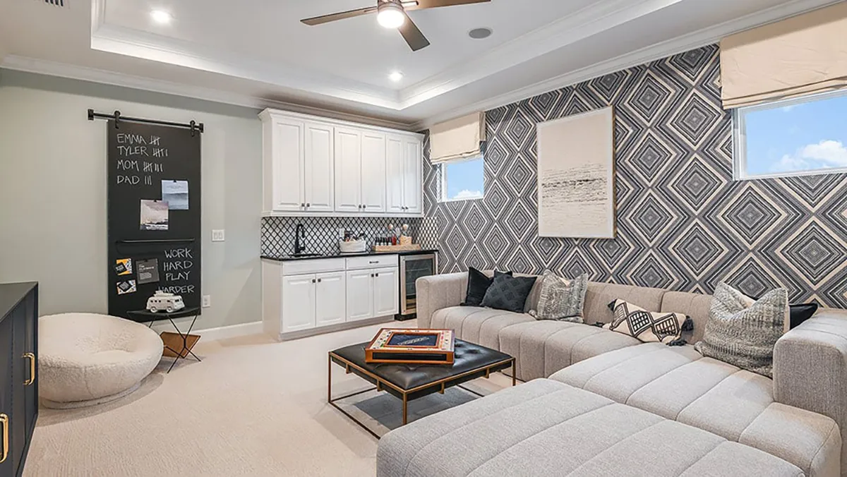 Modern living room with a large sectional sofa, patterned accent wall, mini kitchenette, geometric wallpaper, wall art, a chalkboard with notes, bean bag chair, coffee table, and neutral-toned décor.