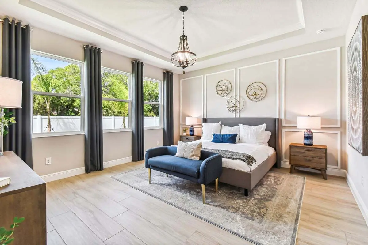 A bright, modern bedroom with a large bed, blue bench, bedside tables with lamps, wall art, three tall windows with dark curtains, and light wood flooring. The room has neutral tones and natural light.