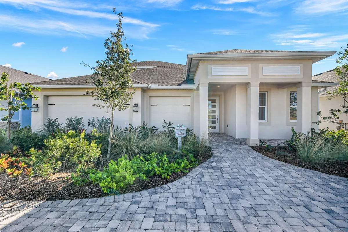 Single-story modern house with a light exterior, large windows, double garage, and a paved walkway leading to the front entrance, surrounded by landscaped greenery under a bright blue sky.