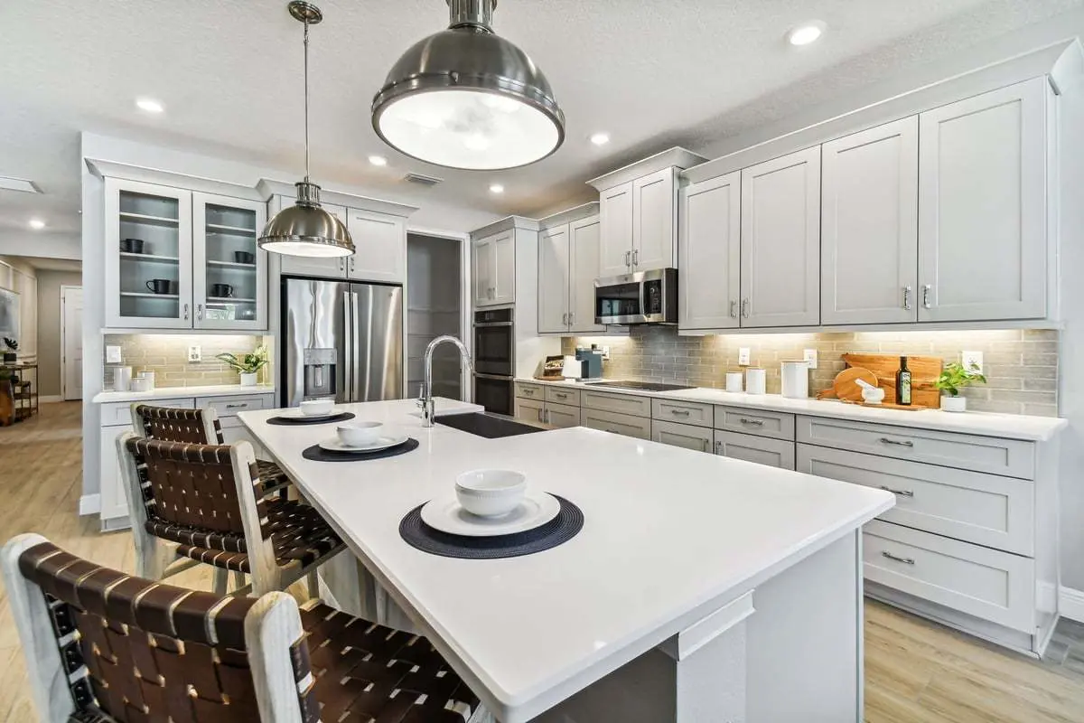 Modern kitchen with white cabinets, stainless steel appliances, an island with two place settings, woven barstools, pendant lights, and light wood flooring. The space is bright and organized with neutral tones.