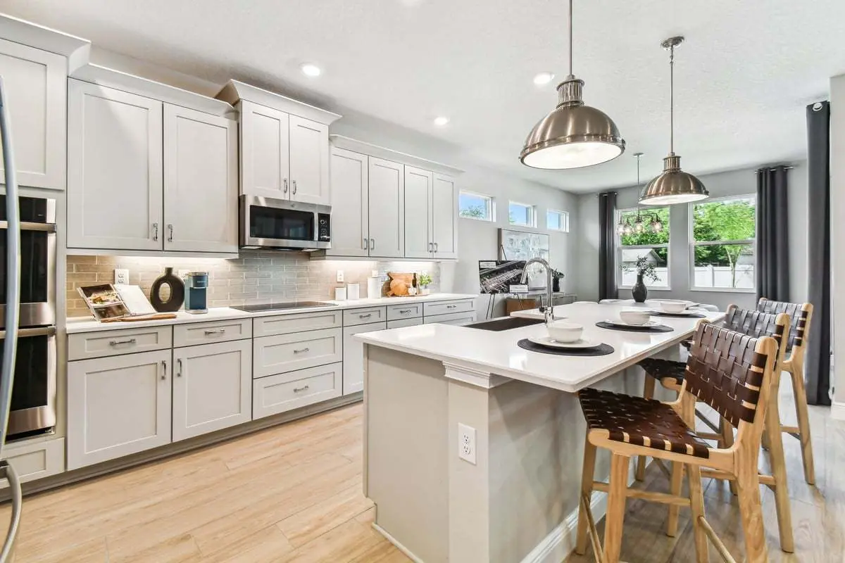 Modern kitchen with white cabinets, stainless steel appliances, a large island with three woven bar stools, pendant lighting, and light wood floors. Large windows let in natural light, creating a bright and airy atmosphere.