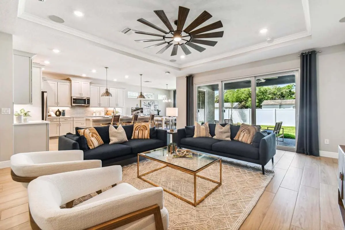 Modern open-concept living room and kitchen with navy sofas, white chairs, a glass coffee table, and a large ceiling fan. Sliding glass doors open to a green backyard. Light wood floors and neutral decor throughout.