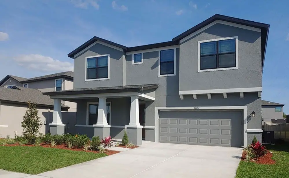 A modern two-story gray house with white trim, a double garage, and a covered front porch. The front yard is landscaped with grass, small plants, and mulch, and the sky is clear and blue.