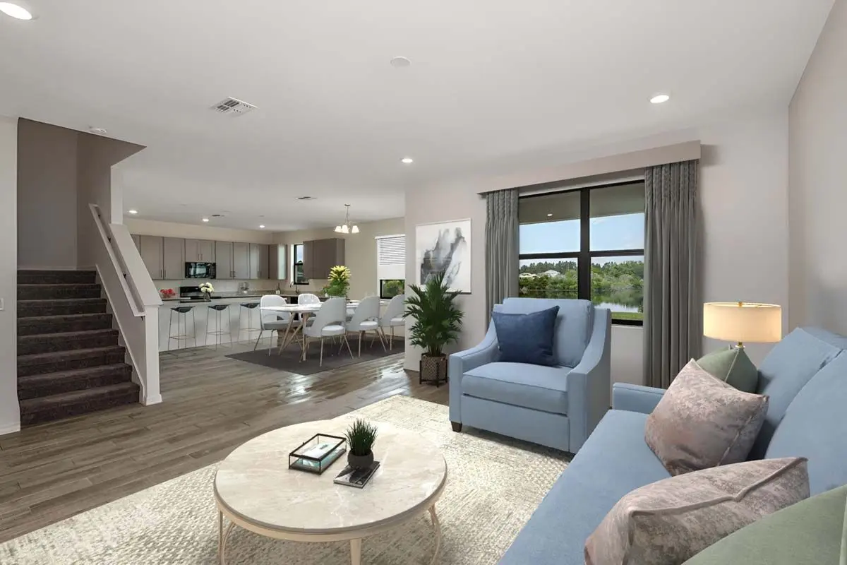 A modern open-plan living room with a blue sofa, armchair, coffee table, and a large window. The space flows into a dining area and kitchen, with stairs leading up on the left and neutral, light décor throughout.