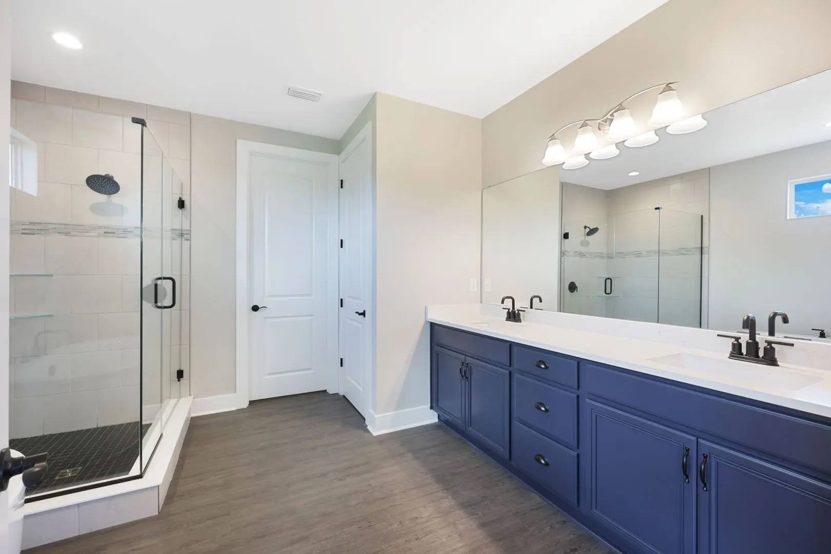 Modern bathroom with a glass-enclosed shower, a double-sink vanity with blue cabinets, large mirror, black fixtures, light wood flooring, and a white door.