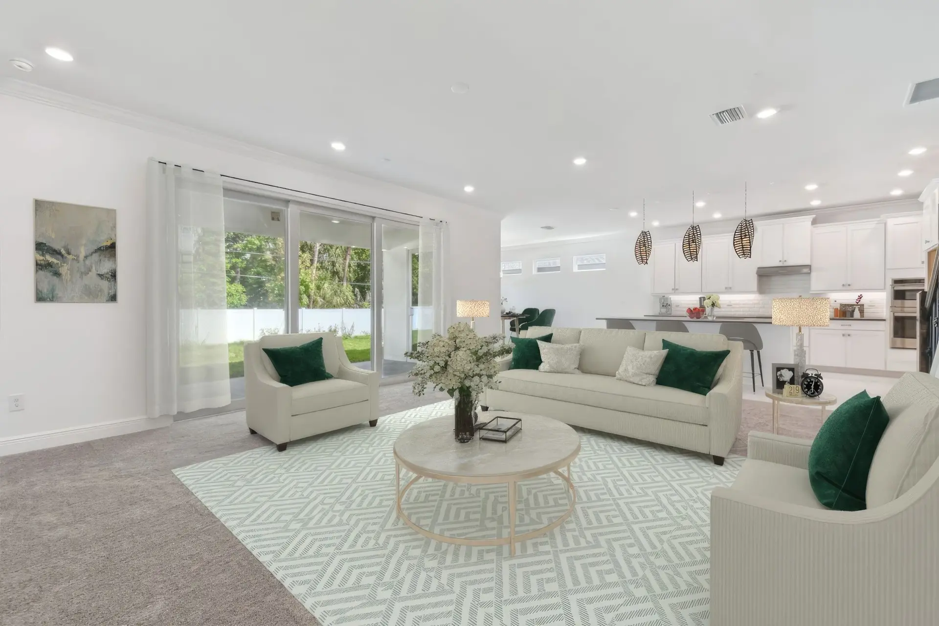A modern living room with beige sofas, green cushions, a round coffee table with flowers, and large glass doors opening to a backyard; the space connects to a kitchen with pendant lights and white cabinetry.