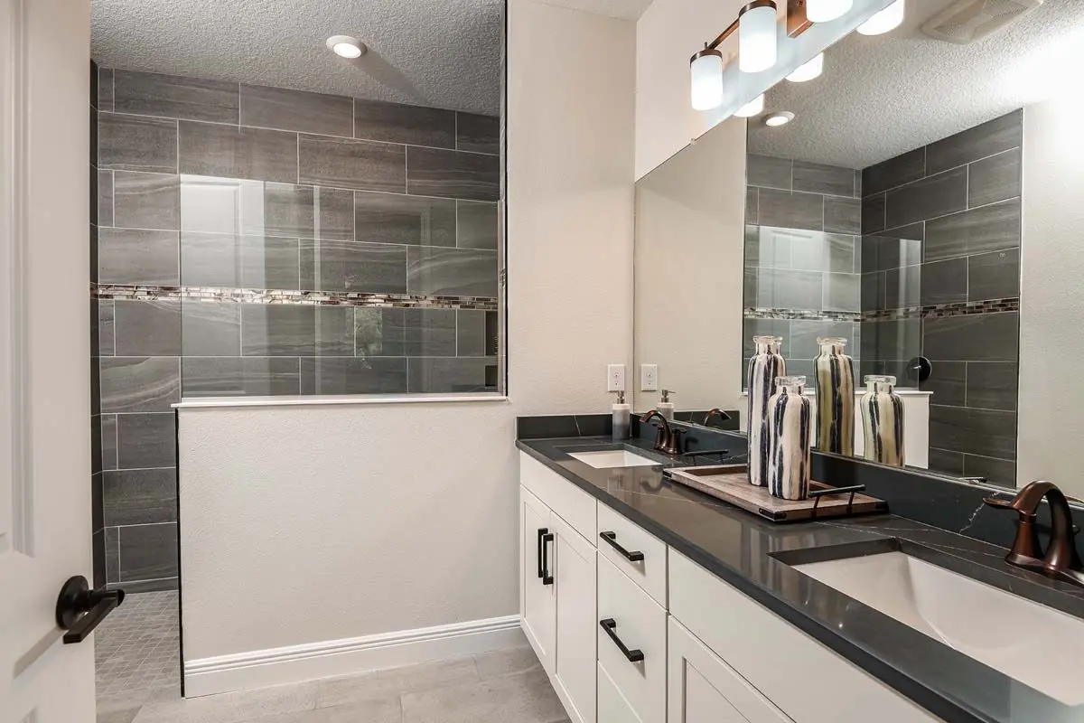 Modern bathroom with a double sink vanity, black countertops, large wall mirror, decorative vases, and a walk-in shower with gray tile and accent strip. Recessed lighting and a three-bulb fixture above the mirror.