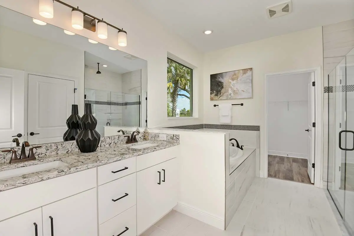 Modern bathroom with a double sink vanity, granite countertop, black decorative vases, built-in bathtub, glass shower, white cabinets, large mirror, wall art, and a window with a view of palm trees.