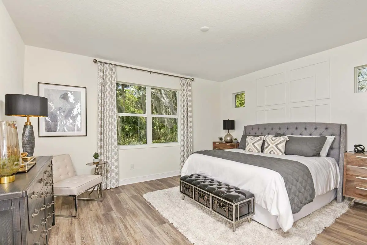 Modern bedroom with a large gray tufted bed, two wooden nightstands, a bench at the foot of the bed, neutral decor, a large window with patterned curtains, and wood flooring.