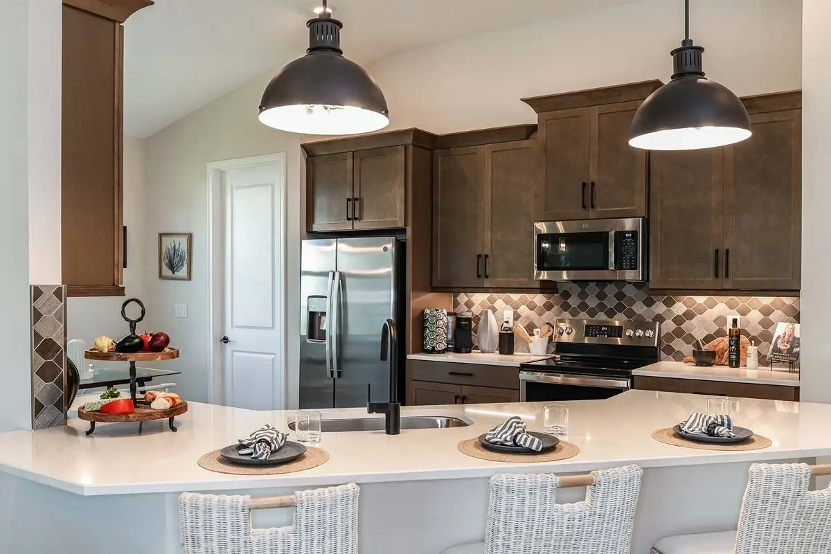Modern kitchen with wood cabinets, stainless steel appliances, and a white island counter set with three place settings. Two large pendant lights hang above, and decorative fruits and kitchen items are visible on the counters.