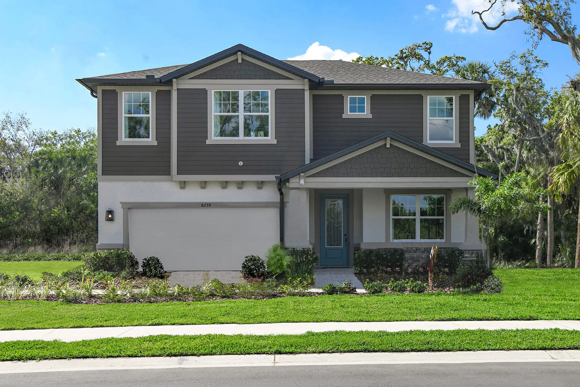 Two-story suburban house with gray and white exterior, double garage, green front lawn, and landscaped shrubs, set against a backdrop of trees and blue sky.