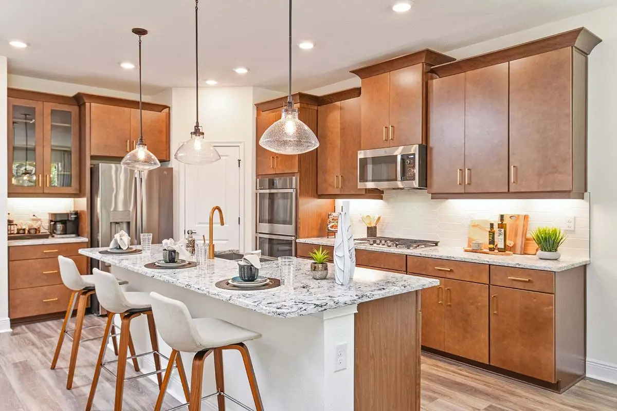 Modern kitchen with wood cabinets, stainless steel appliances, marble island with three white stools, pendant lights, and various decorative items on the counters.