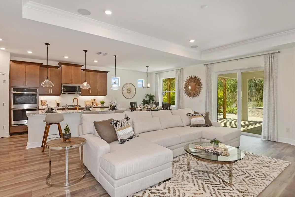 A modern open-concept living room and kitchen with a large beige sectional sofa, round glass coffee table, wooden floors, brown cabinets, pendant lighting, and sliding glass doors opening to a patio and greenery.