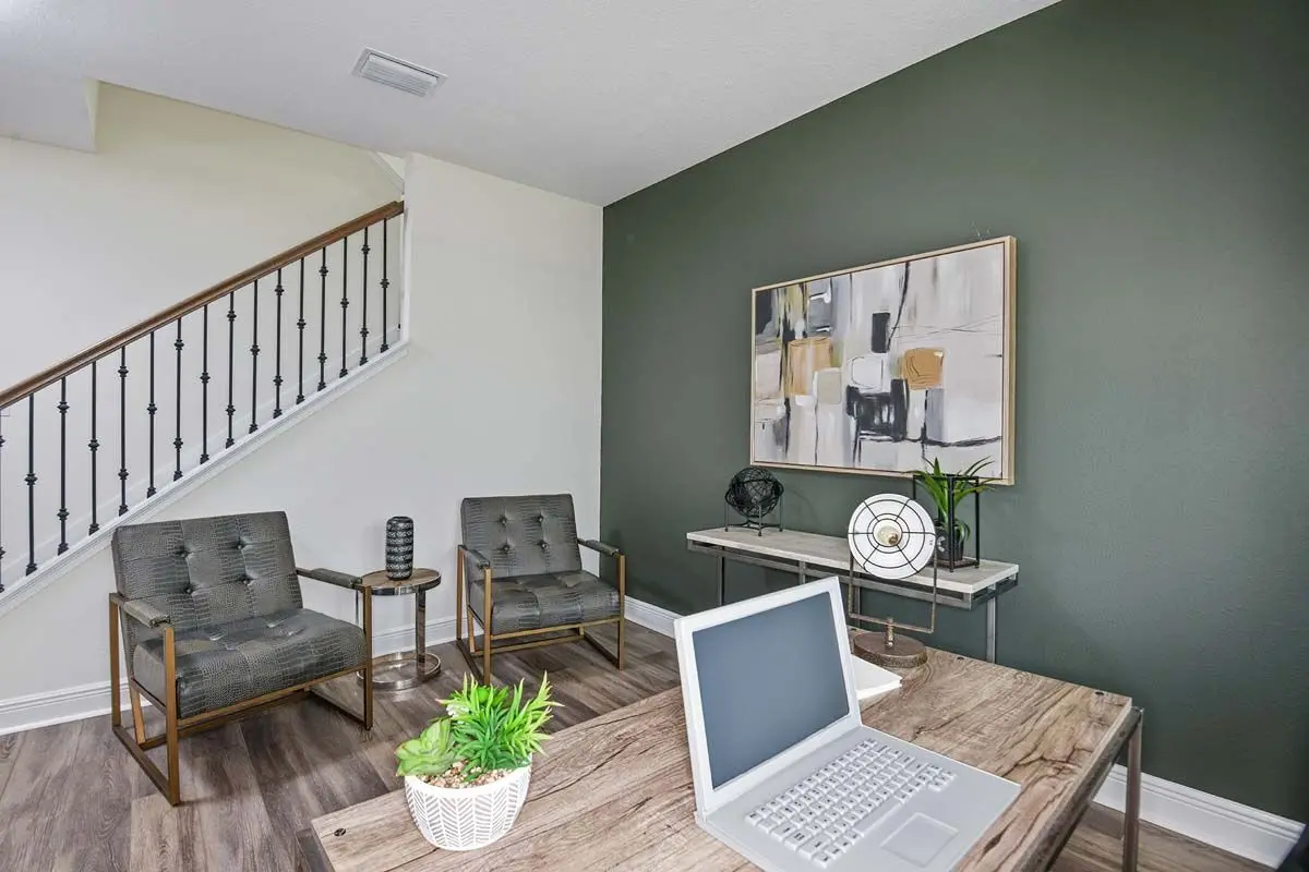 A modern home office with a wooden desk, laptop, potted plant, two gray armchairs, a small side table, abstract wall art, and a staircase with a decorative railing in the background.