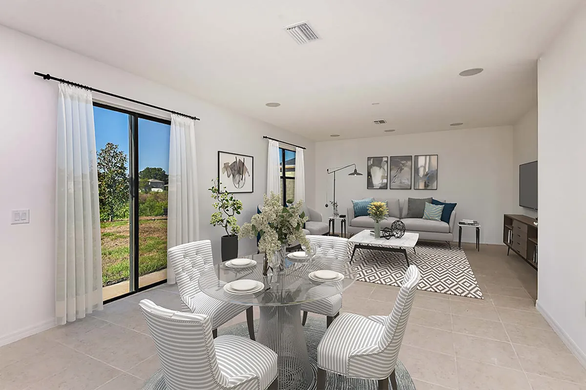 Modern open-plan living and dining area with a round glass table set for four, striped chairs, a white sofa, wall art, and a large window with sheer curtains overlooking greenery.